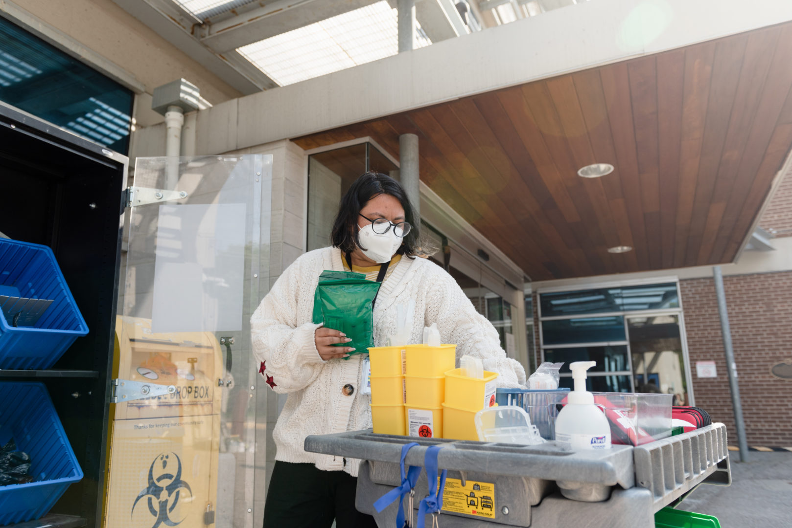 a worker with short black hair and glasses wears a mask and sorts through medical supplies on a cart. They hold small green bags in their hand while they sort.