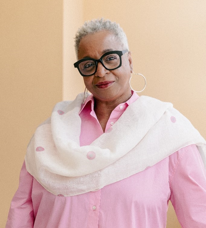 Hzelle palmer, seen from the chest up, wearing a pink collared shirt, large silver hoop earrings , black glasses, and short grey hair
