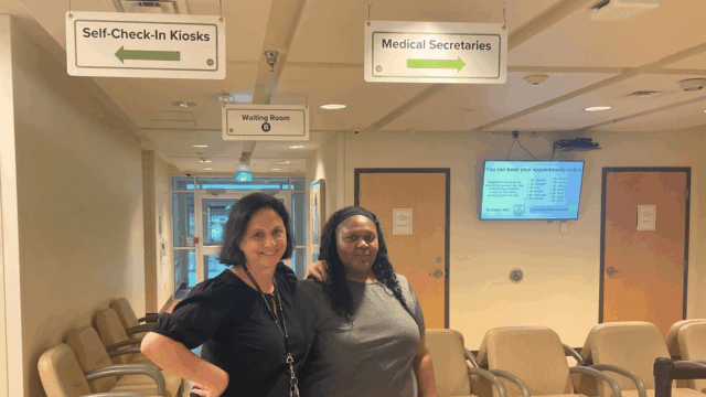 two medical secretaries stand in a waiting room with signs directing to reception and self check-in kiosks