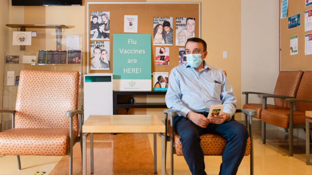 A client sitting in a waiting room at Sherbourne Health Centre. Behind them is a poster sign that reads, 