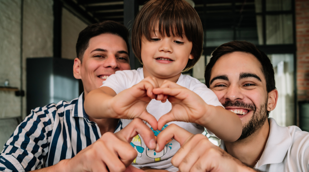 Queer parents and their child making a heart shape with their hands.
