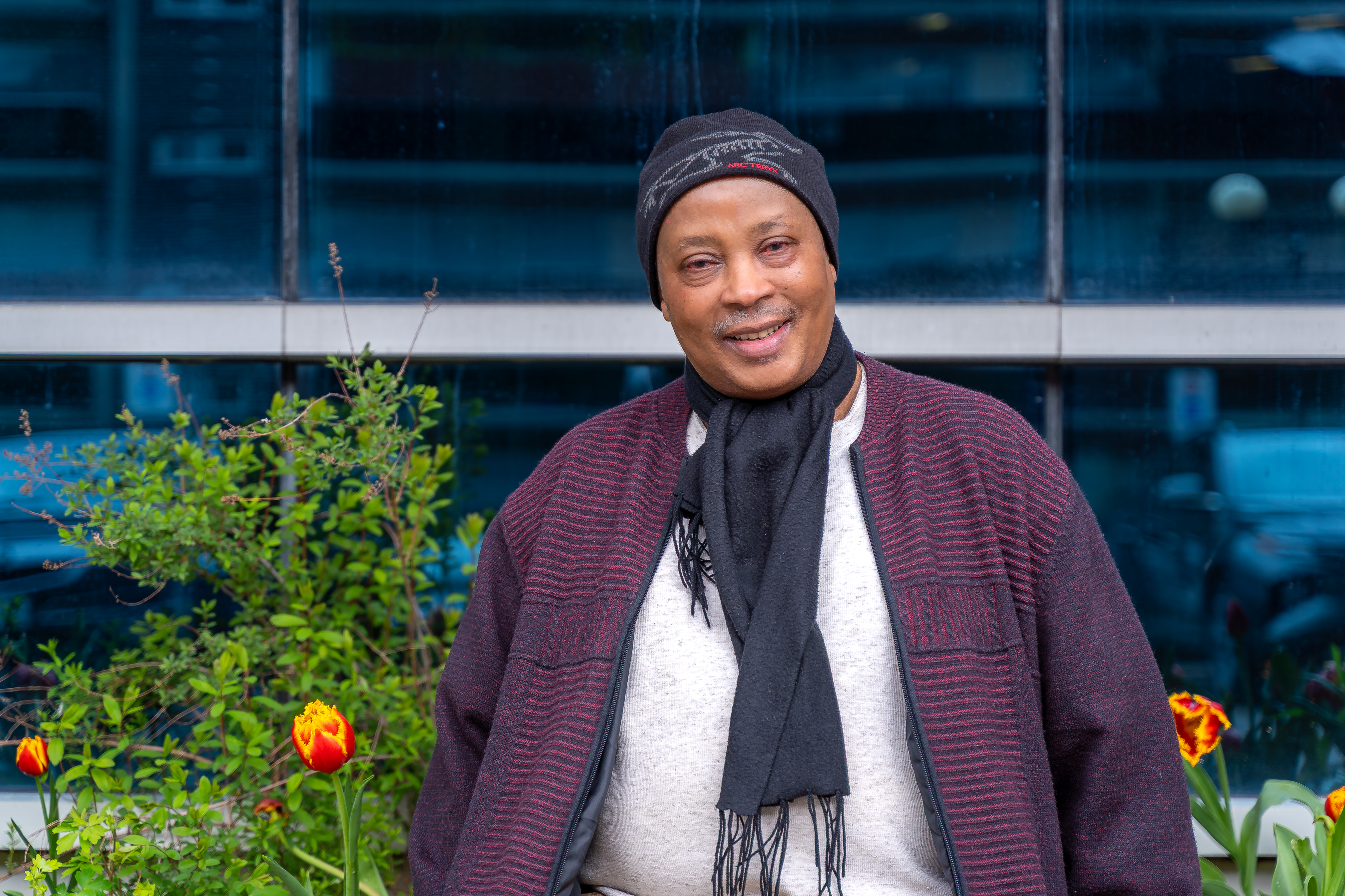 Joseph, wearing a beanie, scarf, and cardigan, poses with a smile outside Sherbourne Health.