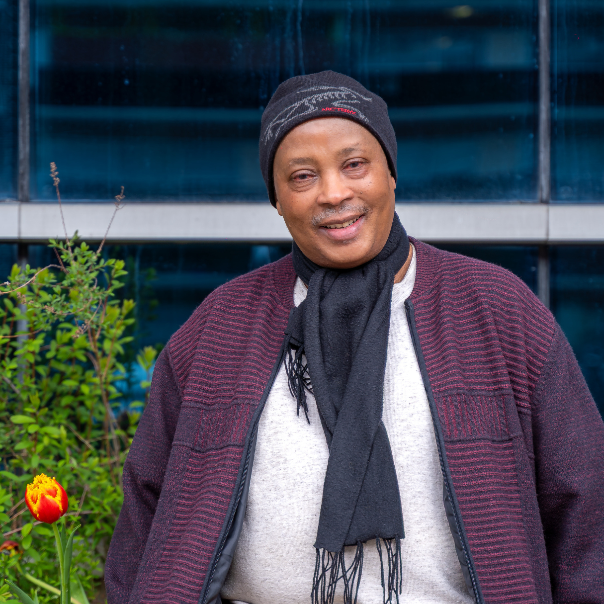 Joseph, wearing a beanie, scarf, and cardigan, poses with a smile outside Sherbourne Health.