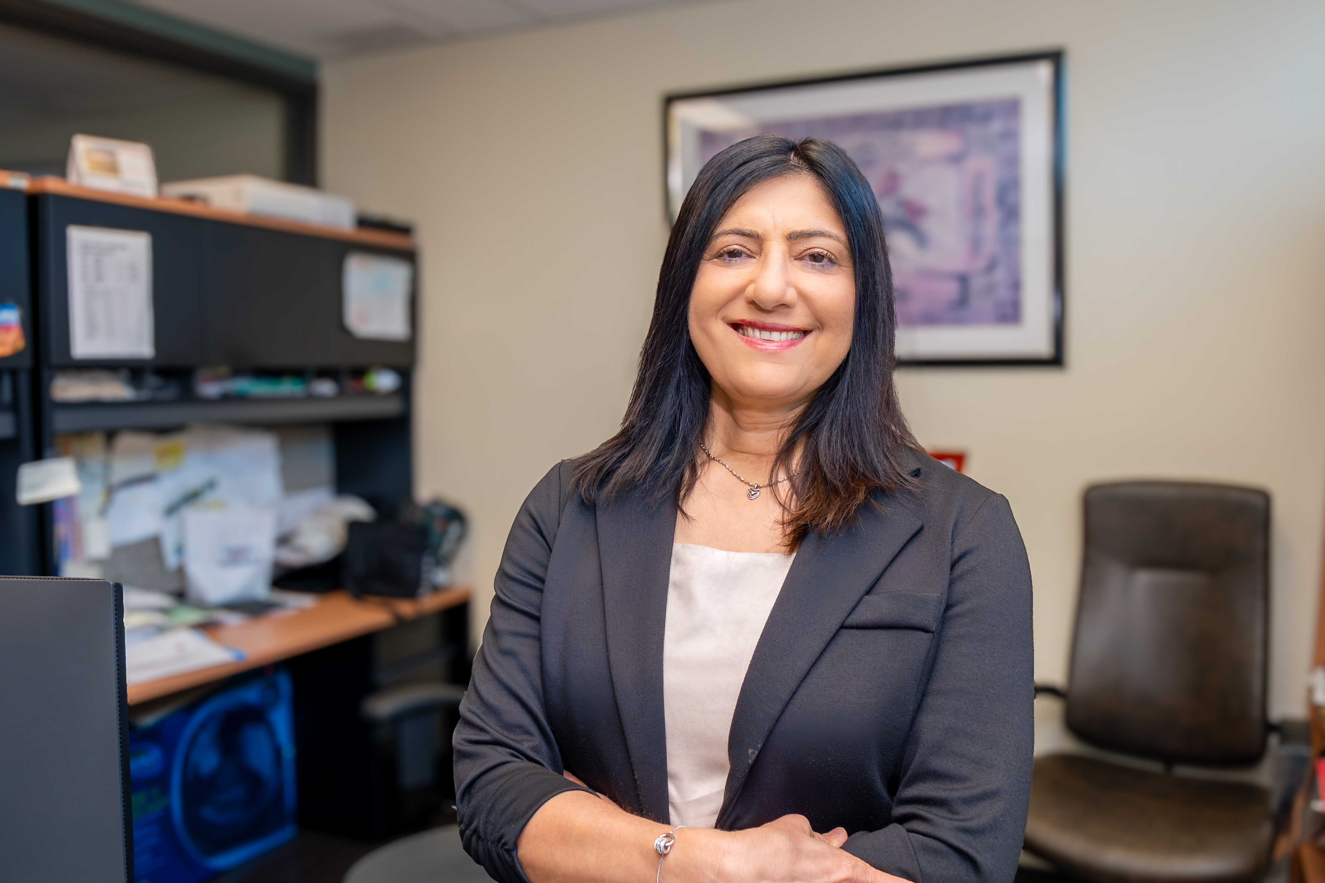 Vinita is a woman with dark shoulder-length hair and dressed in a blazer. She stands in her office, smiling.