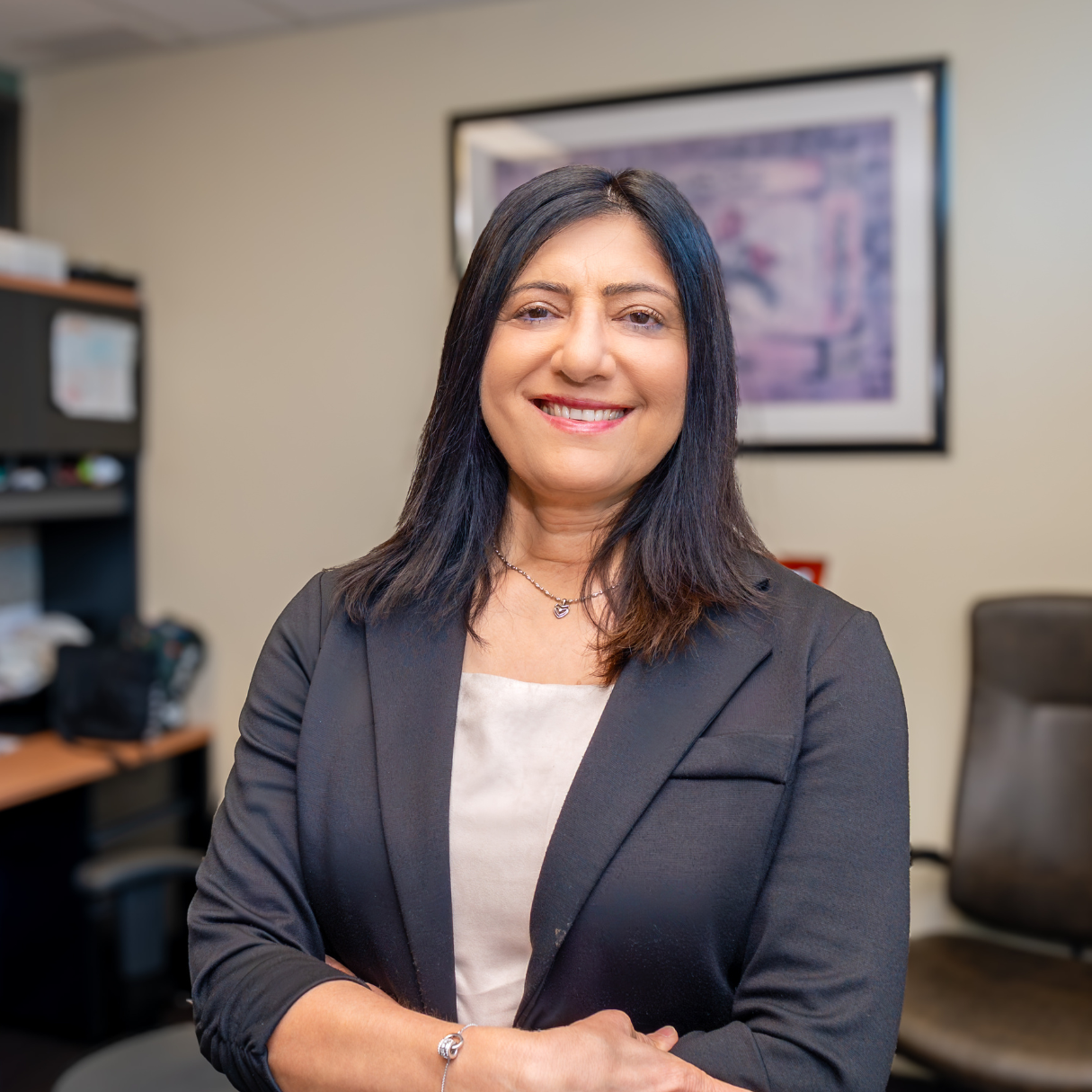 Vinita is a woman with dark shoulder-length hair and dressed in a blazer. She stands in her office, smiling.