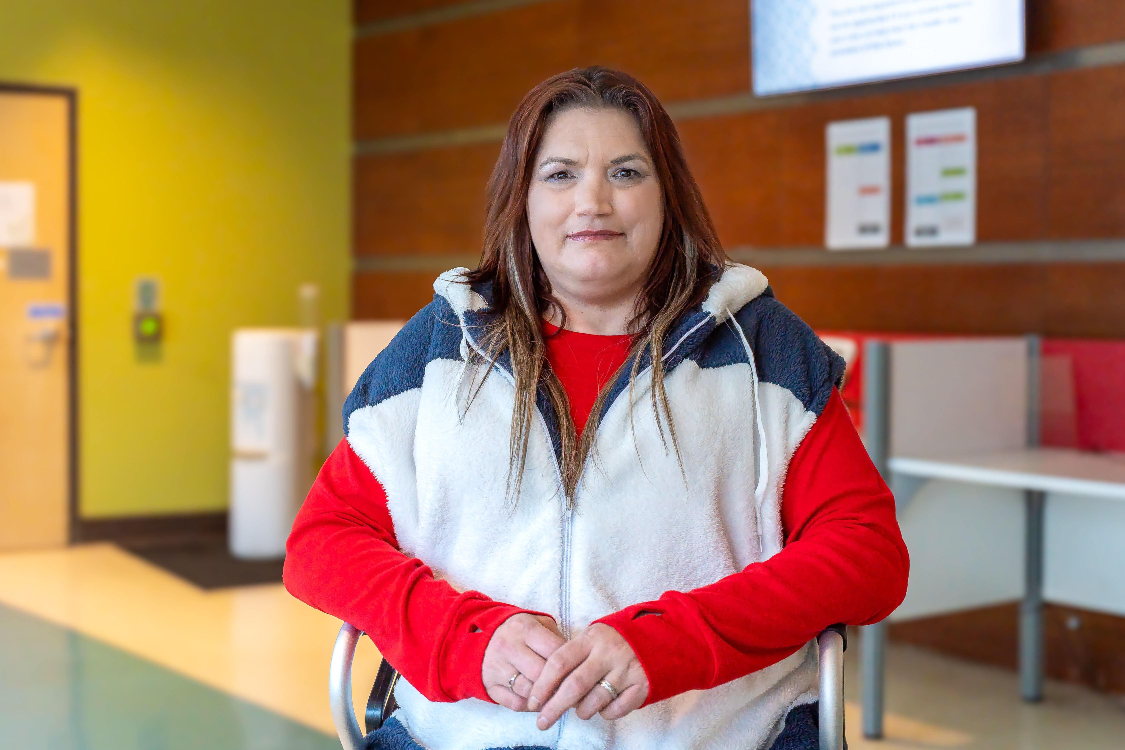 Michelle, a person with below shoulder-length red-brown hair and wearing a red long-sleeved shirt and a white vest jacket, poses inside Sherbourne Health with their hands together.