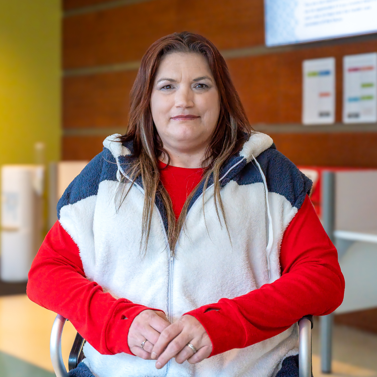 Michelle, a person with below shoulder-length red-brown hair and wearing a red long-sleeved shirt and a white vest jacket, poses inside Sherbourne Health with their hands together.