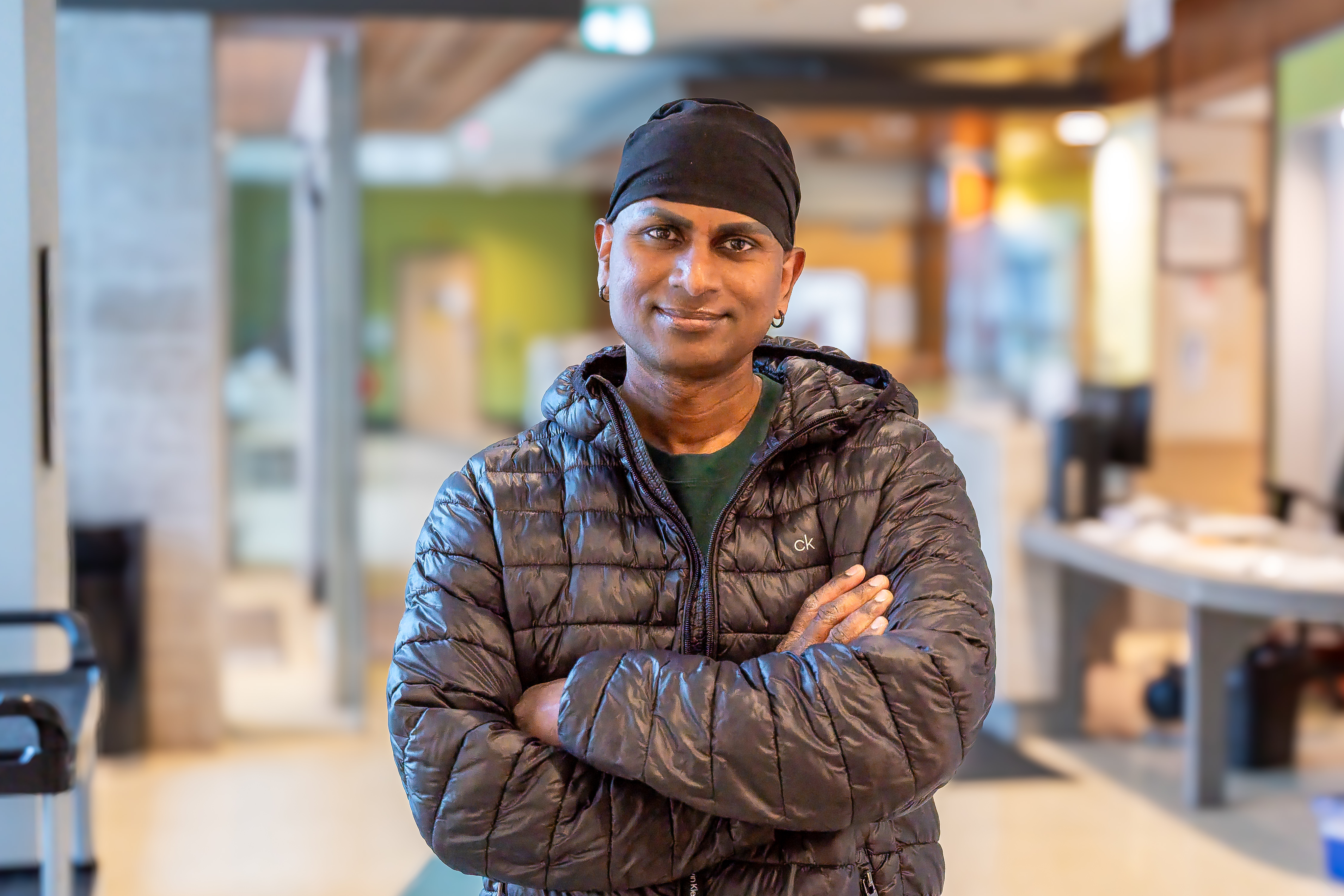 Victor, a client of Sherbourne Health wearing a black jacket and a head covering, stands with his arms crossed in the centre lobby.